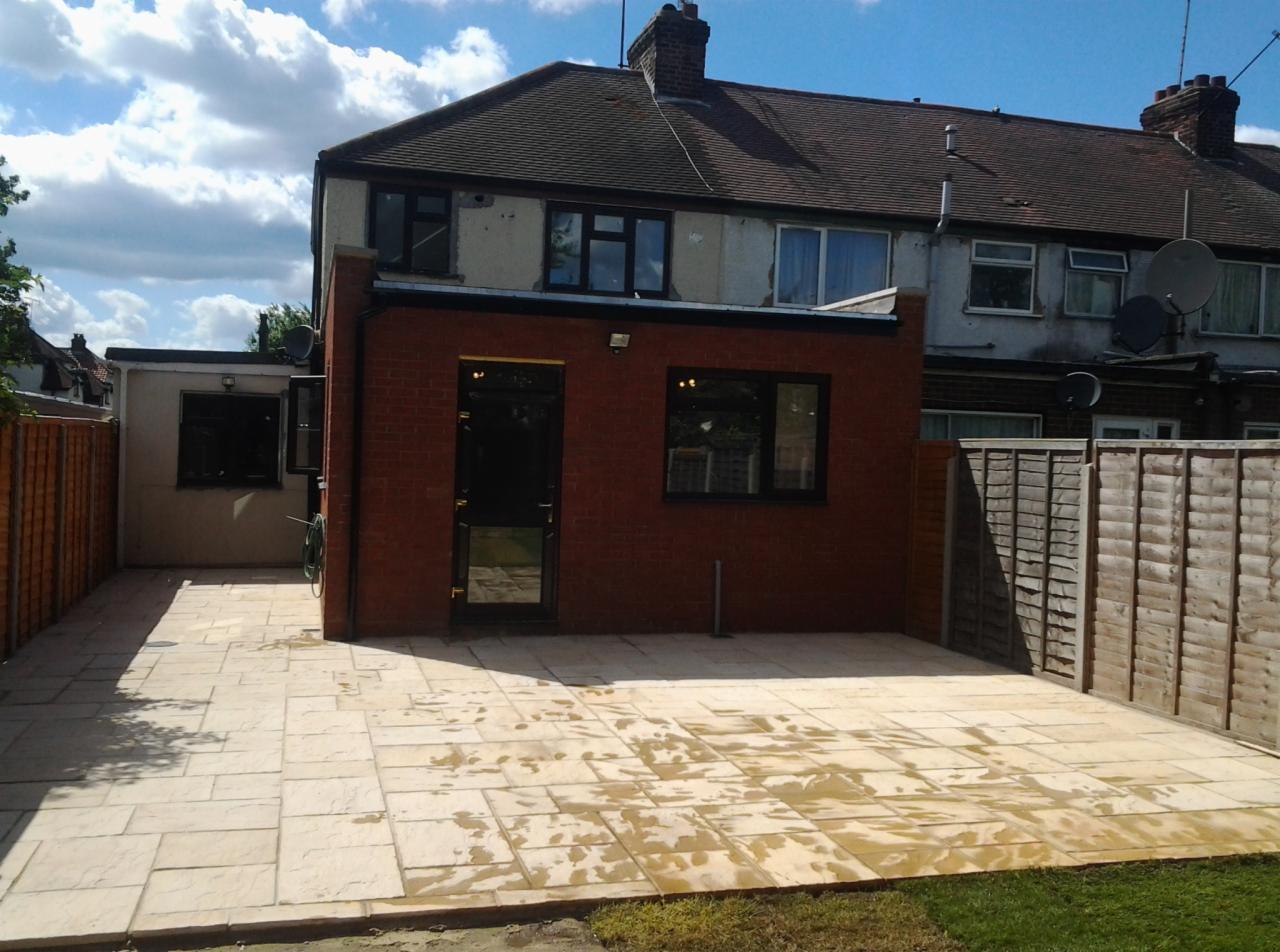 Nowo wyłożony, jasny taras z kostki brukowej przy ceglanym budynku z czarnymi ramami okiennymi i drzwiami, otoczony drewnianym płotem i trawnikiem.