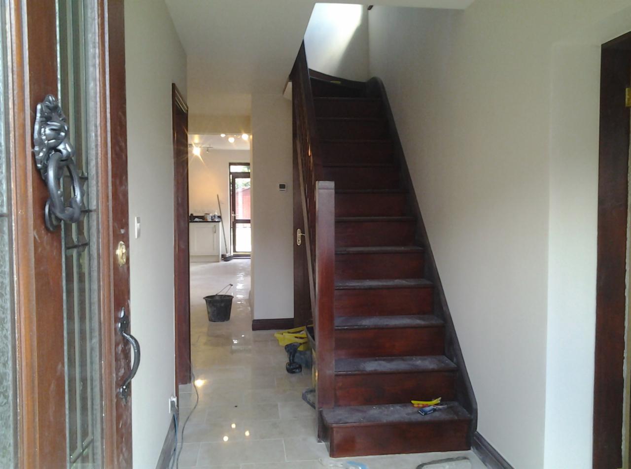 Wnętrze domu w trakcie odświeżania: widok na korytarz z nową podłogą, schody z ciemnego drewna i fragment otwartych drzwi wejściowych z ozdobną kołatką.