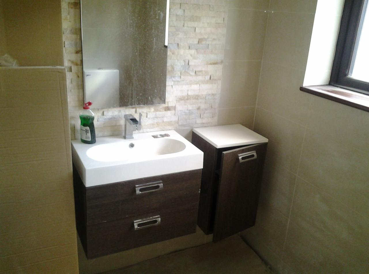 Umywalka z szafką w odcieniach brązu, obok szafka z uchylonymi drzwiczkami, lustro nad umywalką, ściana wyłożona płytkami imitującymi kamień, butelka płynu do mycia na umywalce, widoczne...