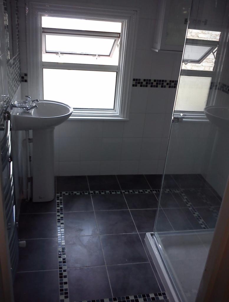 Umywalka na postumencie, kabina prysznicowa ze szklanymi drzwiami i ciemna podłoga z dekoracyjnym obramowaniem w wykończonej łazience z oknem.