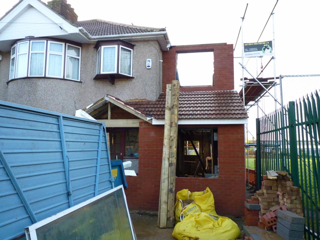 Budowa domu: dobudowa z czerwonej cegły w trakcie realizacji, widoczne rusztowanie, materiały budowlane i fragment istniejącego budynku