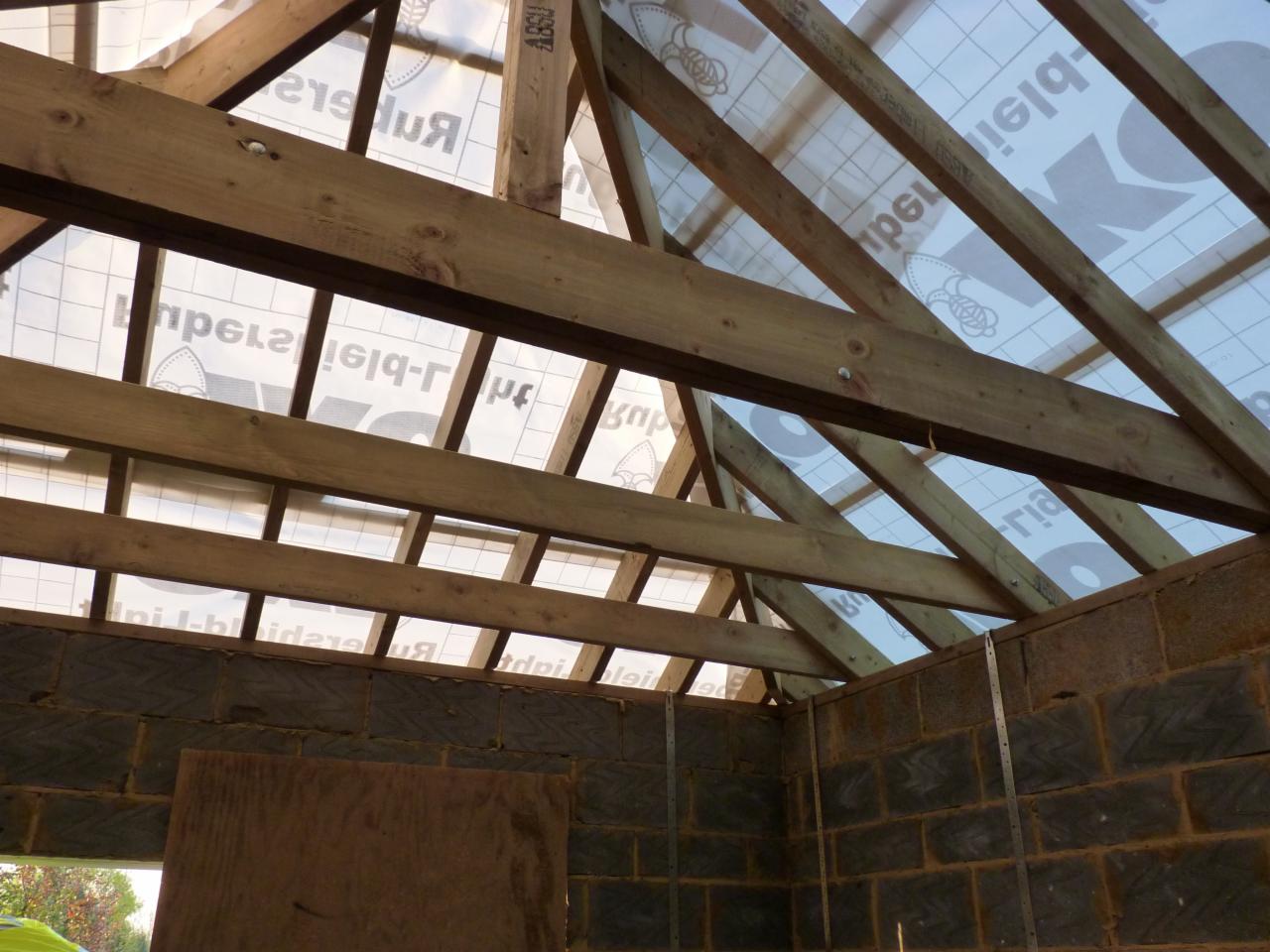 Wnętrze surowego domu w trakcie budowy, widoczne ściany z bloczków betonowych, drewniana konstrukcja dachu z membraną dachową z nadrukiem, metalowe łączniki wzmacniające ściany.