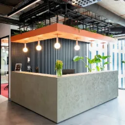 Nowoczesna recepcja w biurze z betonowym blatem, pionowymi czarnymi listwami na ścianie, pomarańczową konstrukcją sufitową z roślinami i trzema wiszącymi lampami kulami, w tle szklane biuro i okna...