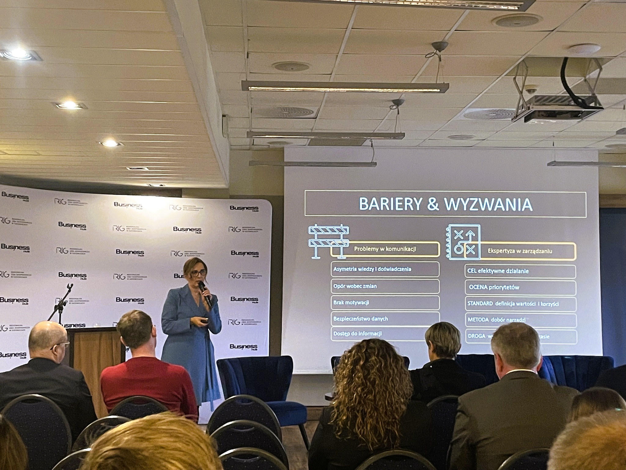 Prelegentka w niebieskiej sukience prezentuje slajd 'Bariery i wyzwania' podczas szkolenia dla firm. Widzowie siedzą na krzesłach, słuchając prelekcji.