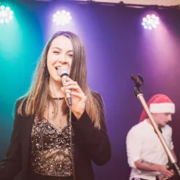 Wokalistka z długimi włosami śpiewa do mikrofonu w błyszczącym topie i czarnej marynarce na tle kolorowych świateł, w tle perkusista w czapce Świętego Mikołaja.