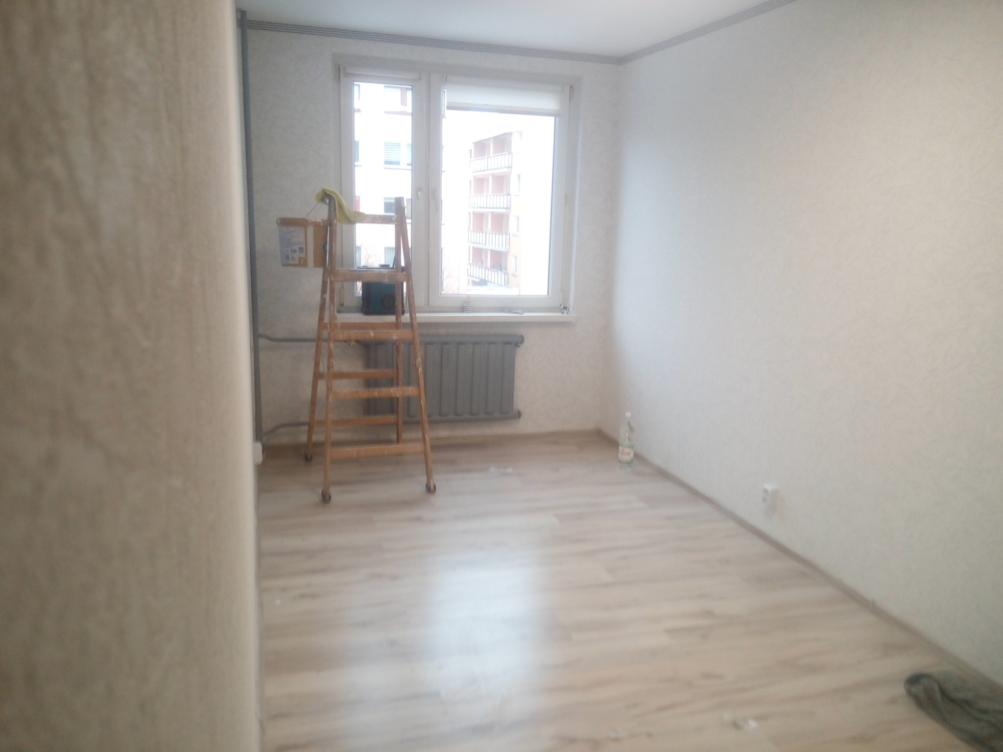 Pokój w trakcie prac wykończeniowych: drabina, wiadro z farbą, okno z widokiem na budynek wielorodzinny, panele podłogowe, ściany przygotowane do malowania.
