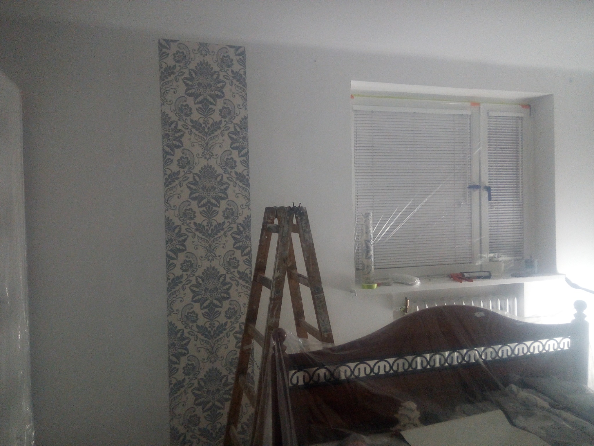 Fragment pokoju w trakcie prac: ściana z tapetą w kwiaty, drewniana drabina malarska, zabezpieczone folią łóżko, okno z żaluzjami.