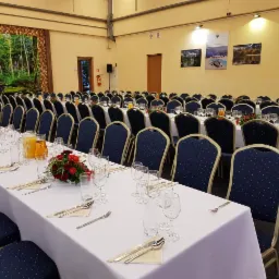 Uroczysta sala bankietowa z długimi stołami nakrytymi białymi obrusami, ustawionymi rzędami, z ciemnoniebieskimi krzesłami w kropki, kieliszkami, sztućcami, serwetkami, dzbankami z sokiem...