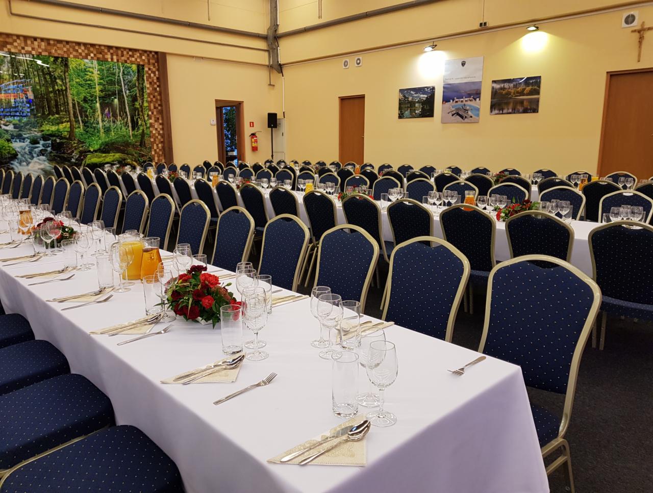 Uroczysta sala bankietowa z długimi stołami nakrytymi białymi obrusami, ustawionymi rzędami, z ciemnoniebieskimi krzesłami w kropki, kieliszkami, sztućcami, serwetkami, dzbankami z sokiem...