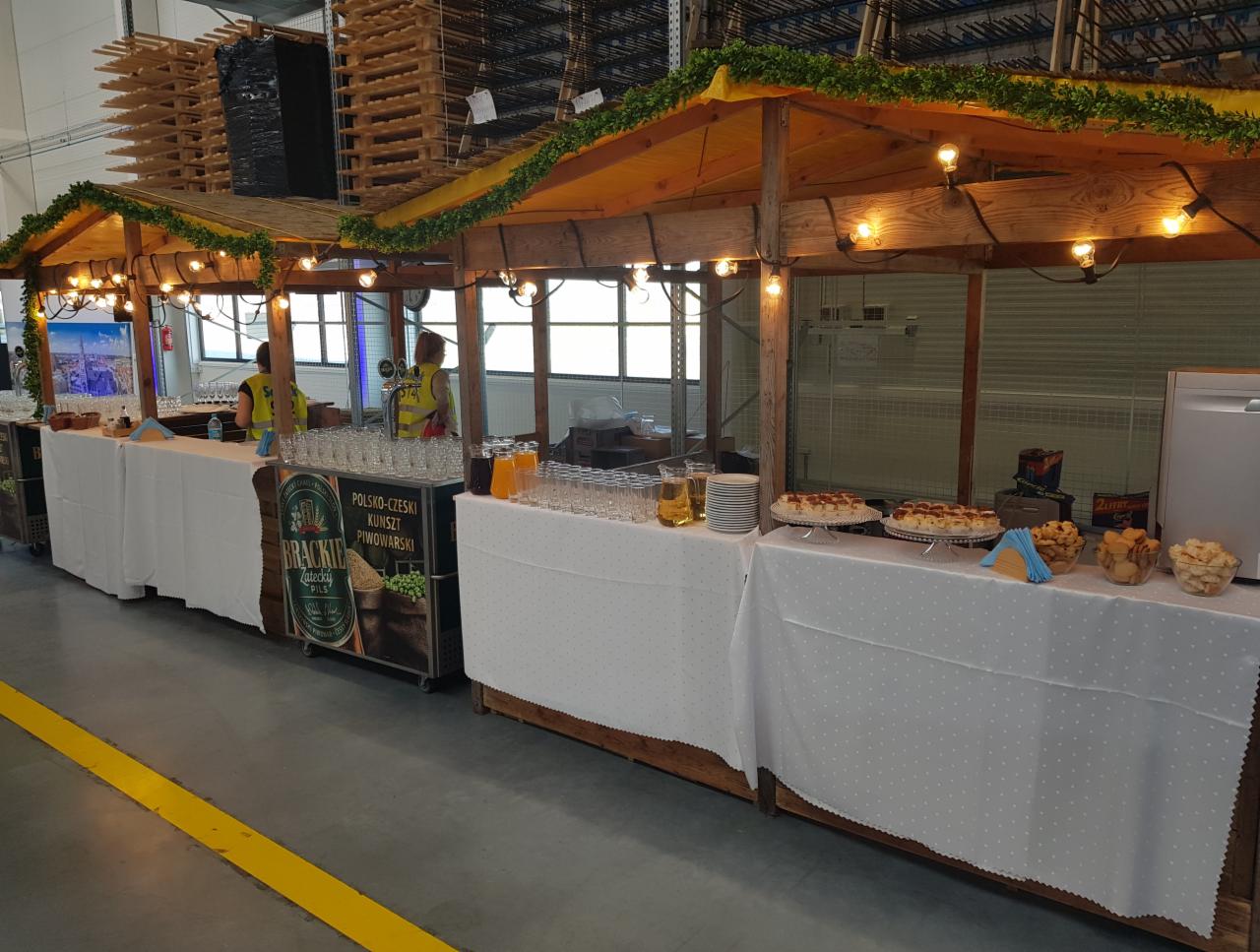 Długi stół cateringowy z drewnianą konstrukcją z oświetleniem, dekorowany girlandą, z napojami, przekąskami i obsługą w tle, ustawiony w hali magazynowej.