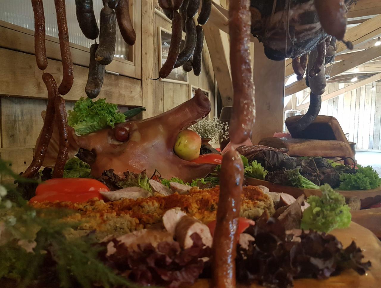 Stół z wędlinami, głową świni z jabłkiem w pysku, wiszącymi kiełbasami i sałatą, w rustykalnej scenerii drewnianej chaty.