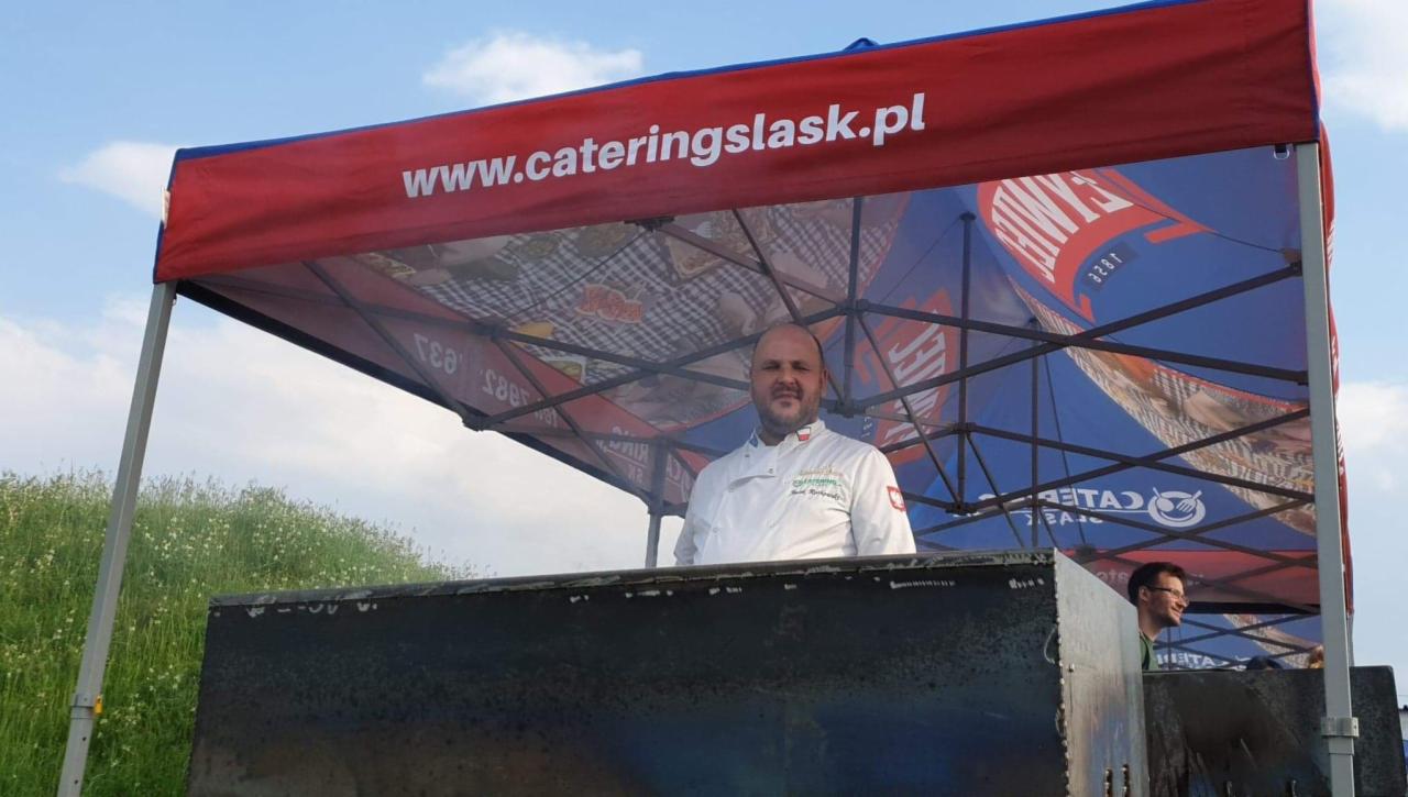 Kucharz w białym uniformie pod namiotem cateringowym z logo www.cateringslask.pl, przygotowujący potrawy na grillu podczas imprezy plenerowej.