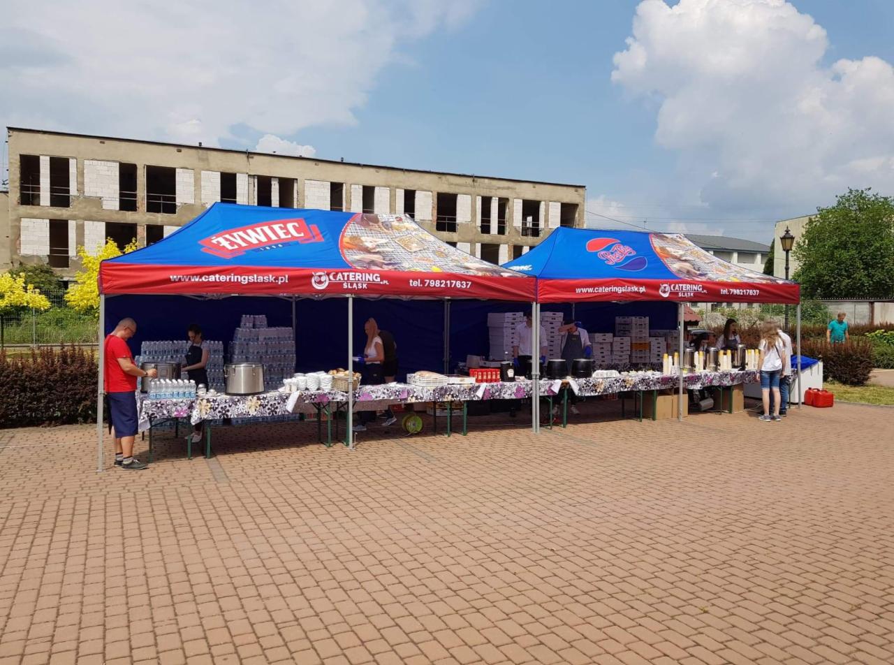 Dwa namioty cateringowe z logo firmy, stoły zastawione jedzeniem i napojami, obsługa w tle, budynek w tle, dzień słoneczny.