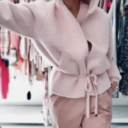 Uśmiechnięta kobieta w różowym komplecie: futrzana bluza z kapturem i spodnie. Okulary przeciwsłoneczne odbijają otoczenie. W tle wieszaki z ubraniami.