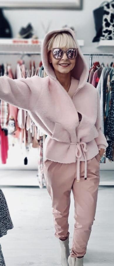 Uśmiechnięta kobieta w różowym komplecie: futrzana bluza z kapturem i spodnie. Okulary przeciwsłoneczne odbijają otoczenie. W tle wieszaki z ubraniami.