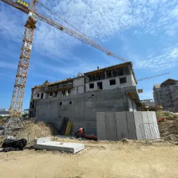 Wysoki, surowy budynek w trakcie budowy z betonu i cegły, z żółtym żurawiem wieżowym na tle błękitnego nieba z obłokami, widoczni pracownicy na górnych kondygnacjach.