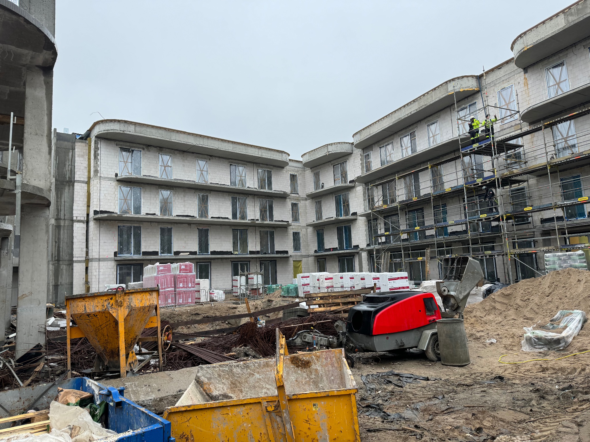 Wielopiętrowy budynek w trakcie wznoszenia z białego bloczka, z widocznymi rusztowaniami, oknami zabezpieczonymi folią, stertami materiałów budowlanych i sprzętem budowlanym na placu budowy...