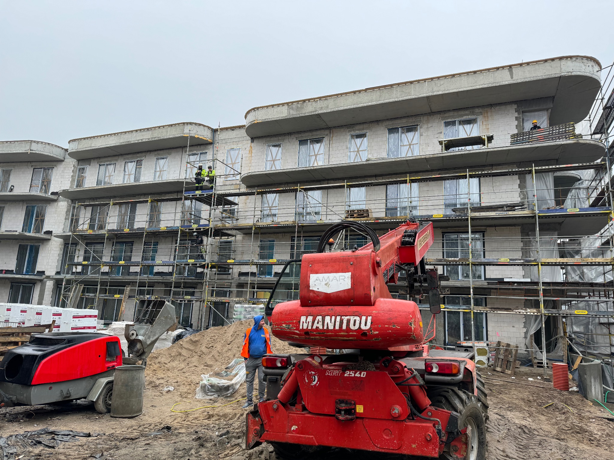 Budowa wielorodzinnego budynku z białego bloczka, z użyciem rusztowań i czerwonego podnośnika teleskopowego Manitou, dwóch robotników na rusztowaniu i jednego przy maszynie.