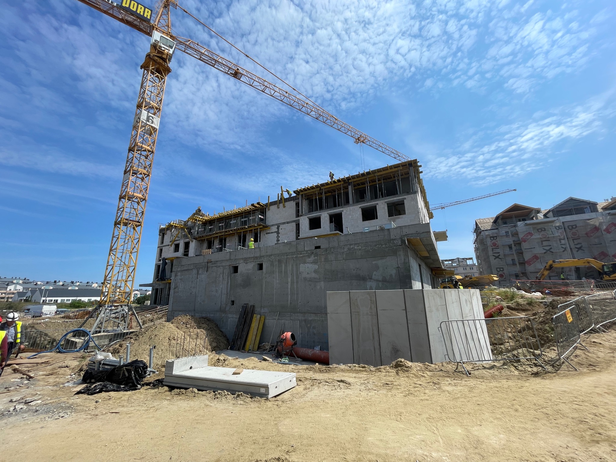 Wysoki, surowy budynek w trakcie budowy z betonu i cegły, z żółtym żurawiem wieżowym na tle błękitnego nieba z obłokami, widoczni pracownicy na górnych kondygnacjach.