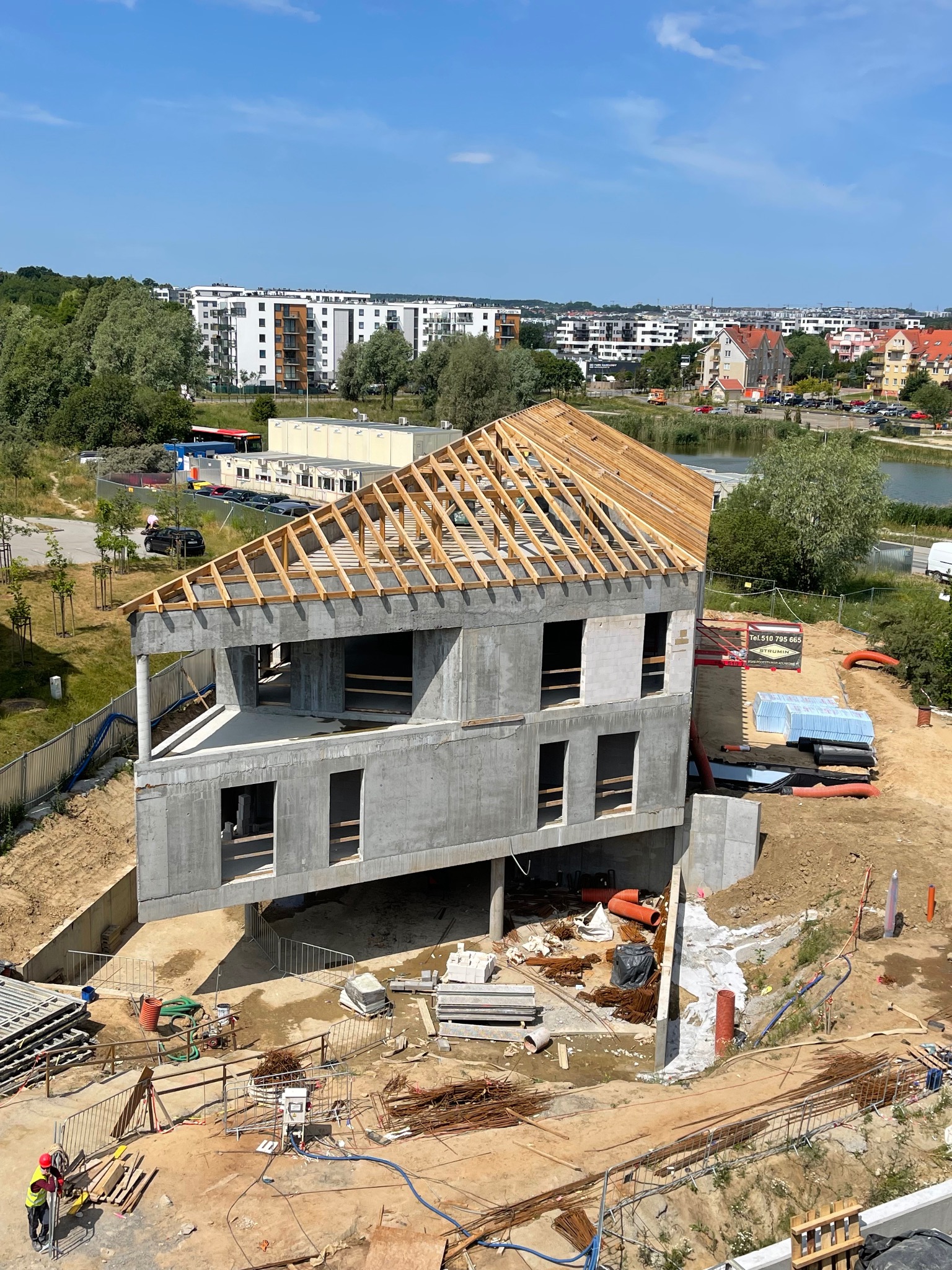 Surowy, dwukondygnacyjny budynek w trakcie budowy z drewnianą konstrukcją dachu, widoczny z góry na tle miejskiego krajobrazu z jeziorem i budynkami mieszkalnymi.