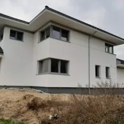 Nowoczesny dom jednorodzinny z białą elewacją i szarymi ramami okiennymi, widoczny z zewnątrz na tle pochmurnego nieba, teren wokół domu częściowo niezagospodarowany z widoczną ziemią i suchą...