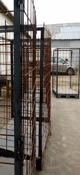 Metalowe klatki spawalnicze ustawione na zewnątrz, widoczne ślady rdzy na niektórych elementach, w tle budynek przemysłowy i zaparkowany samochód.