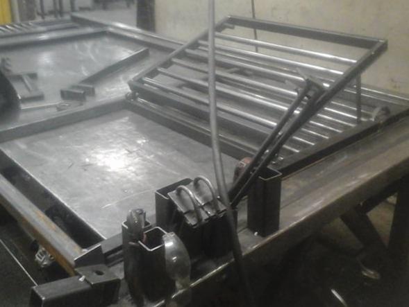 Metalowa konstrukcja spawana z profili i prętów na warsztatowym stole, widoczne narzędzia i elementy do spawania