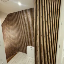 Ściana i schody wykończone panelami dekoracyjnymi z drewna i czarnego filcu, widoczne białe panele podłogowe i biała szafka.