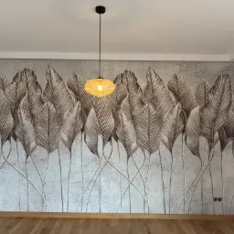 Ściana po tapetowaniu z motywem brązowych liści na szarym tle, oświetlona wiszącą lampą z plecionki, widoczne gniazdka elektryczne i drewniana podłoga.