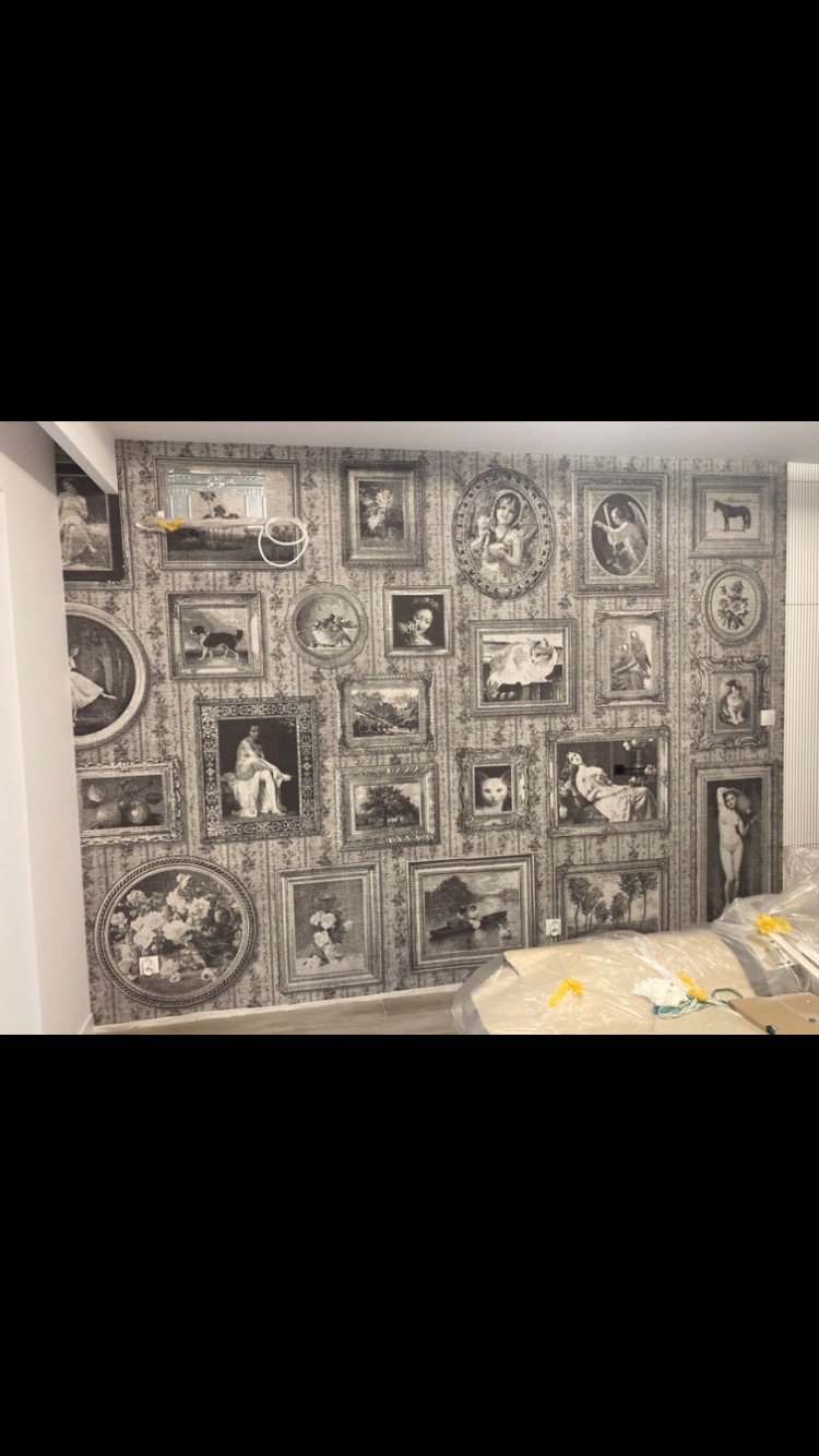 Ściana pokryta tapetą w stylu galerii, z czarno-białymi motywami obrazów w ozdobnych ramach, widoczne zabezpieczone meble w dolnej części kadru.