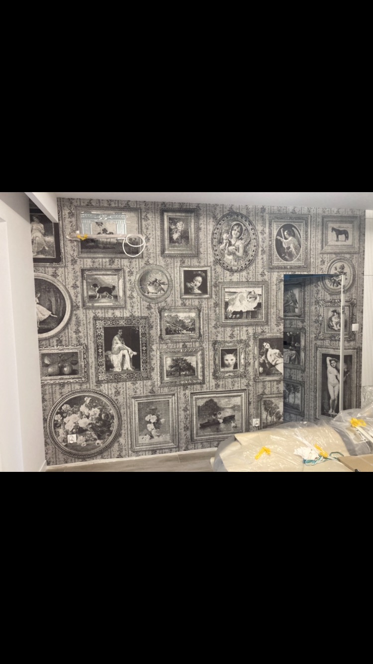 Ściana pokryta tapetą w stylu retro z motywem ramek i obrazów w odcieniach szarości, widoczne niedoskonałości montażu w górnej części ściany i folia ochronna na podłodze.