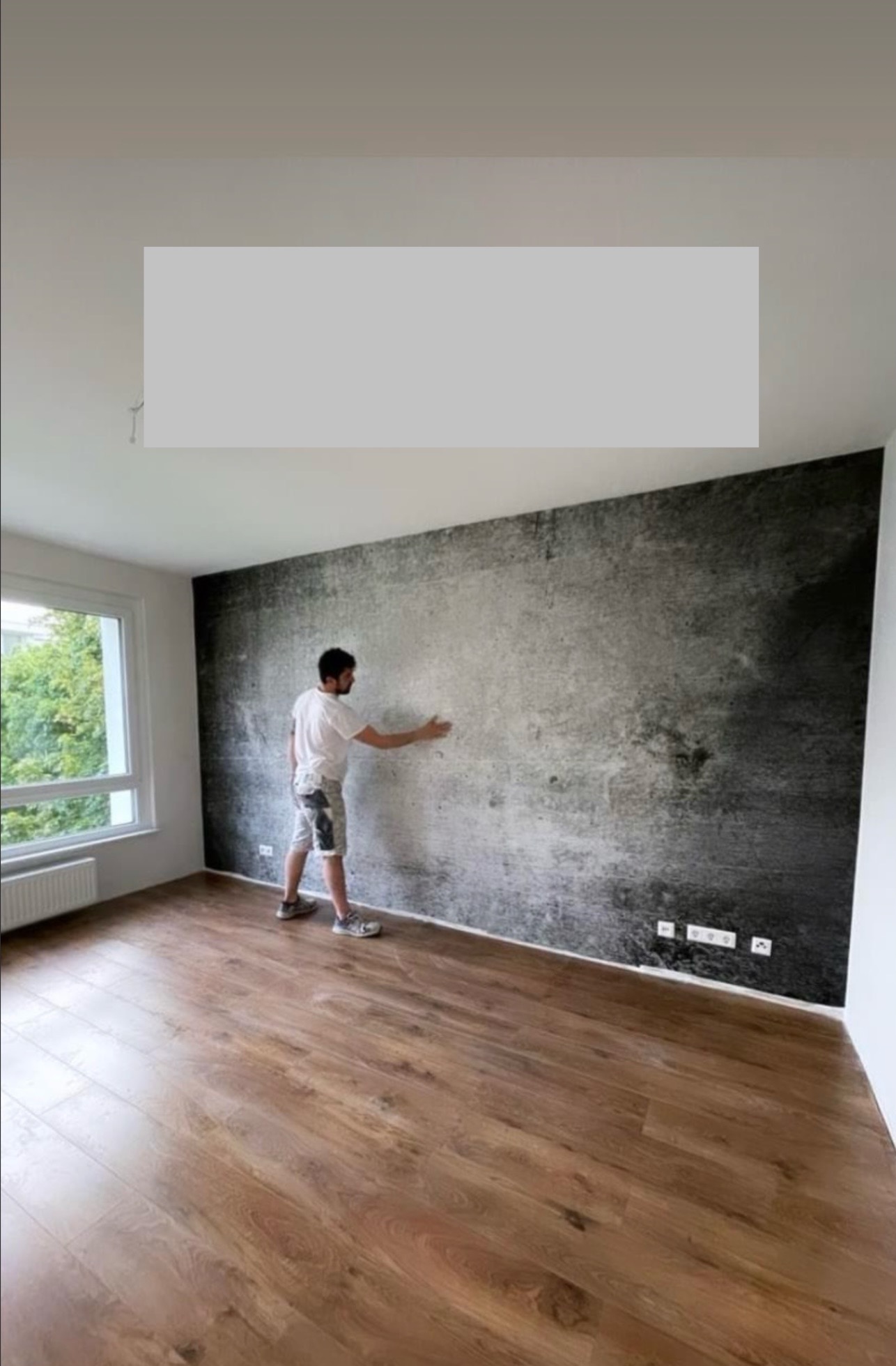 Mężczyzna w spodenkach z plamami farby wygładza dłonią fragment ściany pokrytej tapetą imitującą beton w surowym, minimalistycznym wnętrzu z drewnianą podłogą i oknem z widokiem na zieleń.