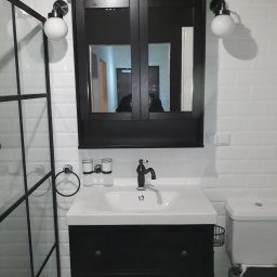 TytusBud - Nowoczesna łazienka z czarną szafką pod umywalką, białymi płytkami i czarną armaturą. W lustrze odbija się osoba. Widoczna kabina prysznicowa z czarnymi ramami.