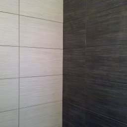 TytusBud - Ściana wyłożona jasnymi i ciemnymi, drewnopodobnymi płytkami ceramicznymi, tworzącymi kontrastowy wzór. Widoczny fragment ościeżnicy drzwi.