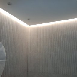 TytusBud - Nowoczesna łazienka z podświetlanym sufitem LED i panelami ściennymi w odcieniach szarości. Minimalistyczny design, widoczny fragment armatury.