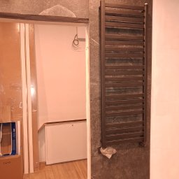 TytusBud - Zamontowany, brązowy grzejnik drabinkowy na szarej, kamiennej ścianie w łazience w trakcie remontu. Widoczne niedokończone wykończenia i materiały budowlane.