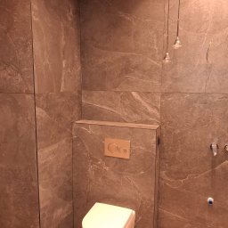 TytusBud - Nowoczesna łazienka z brązowymi płytkami imitującymi kamień, podwieszana toaleta i minimalistyczne oświetlenie. Widoczne detale wykończenia i armatura.