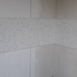 TytusBud - Fragment ściany z mozaiką heksagonalną w odcieniach bieli i szarości, świeżo położoną nad białymi płytkami, widoczne gniazdka elektryczne i rury kanalizacyjne w dolnym rogu.