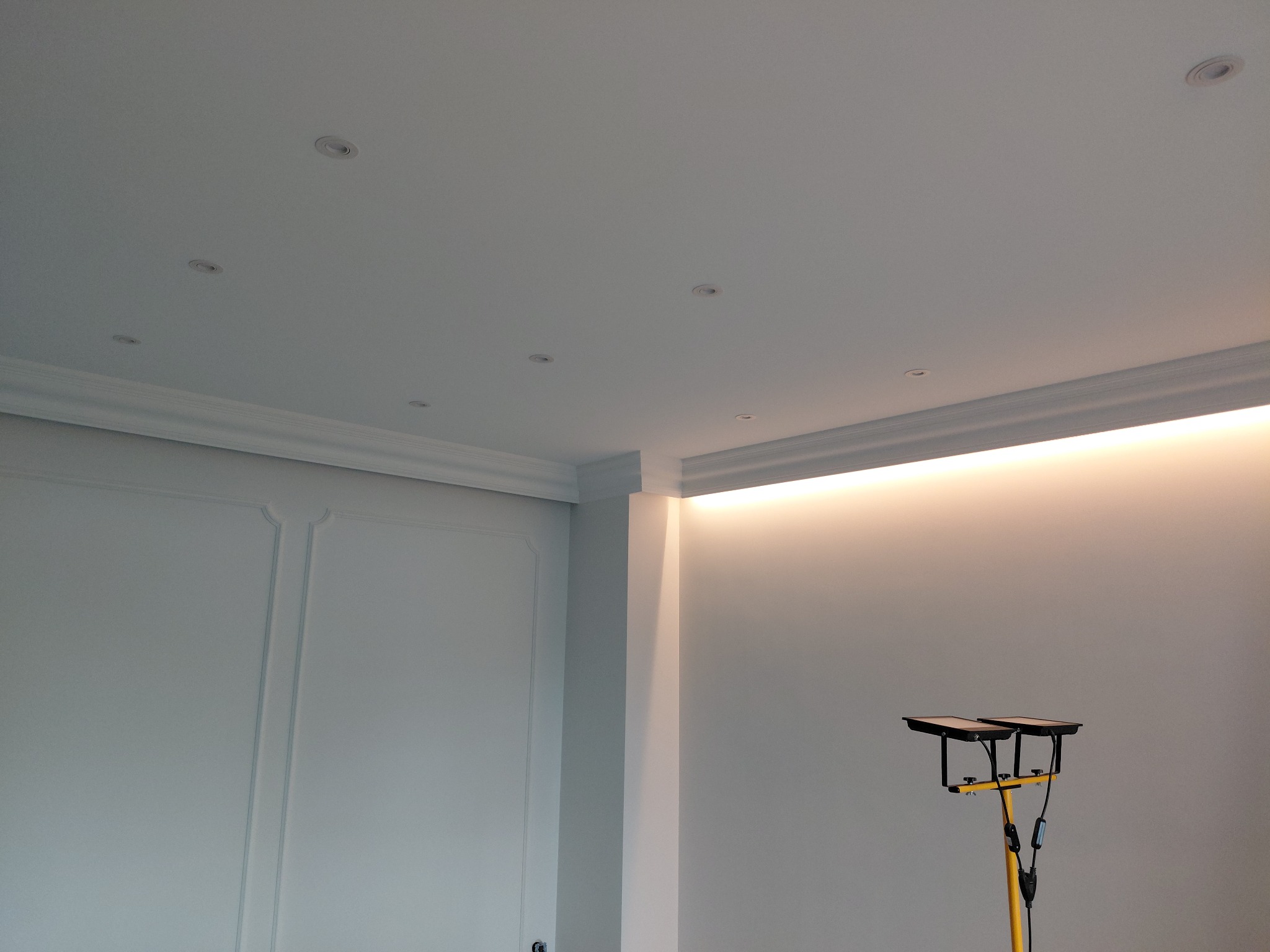 Białe wnętrze po remoncie: ściany z dekoracyjnymi panelami, sufit z oświetleniem punktowym i listwami LED. Na podłodze lampa robocza.