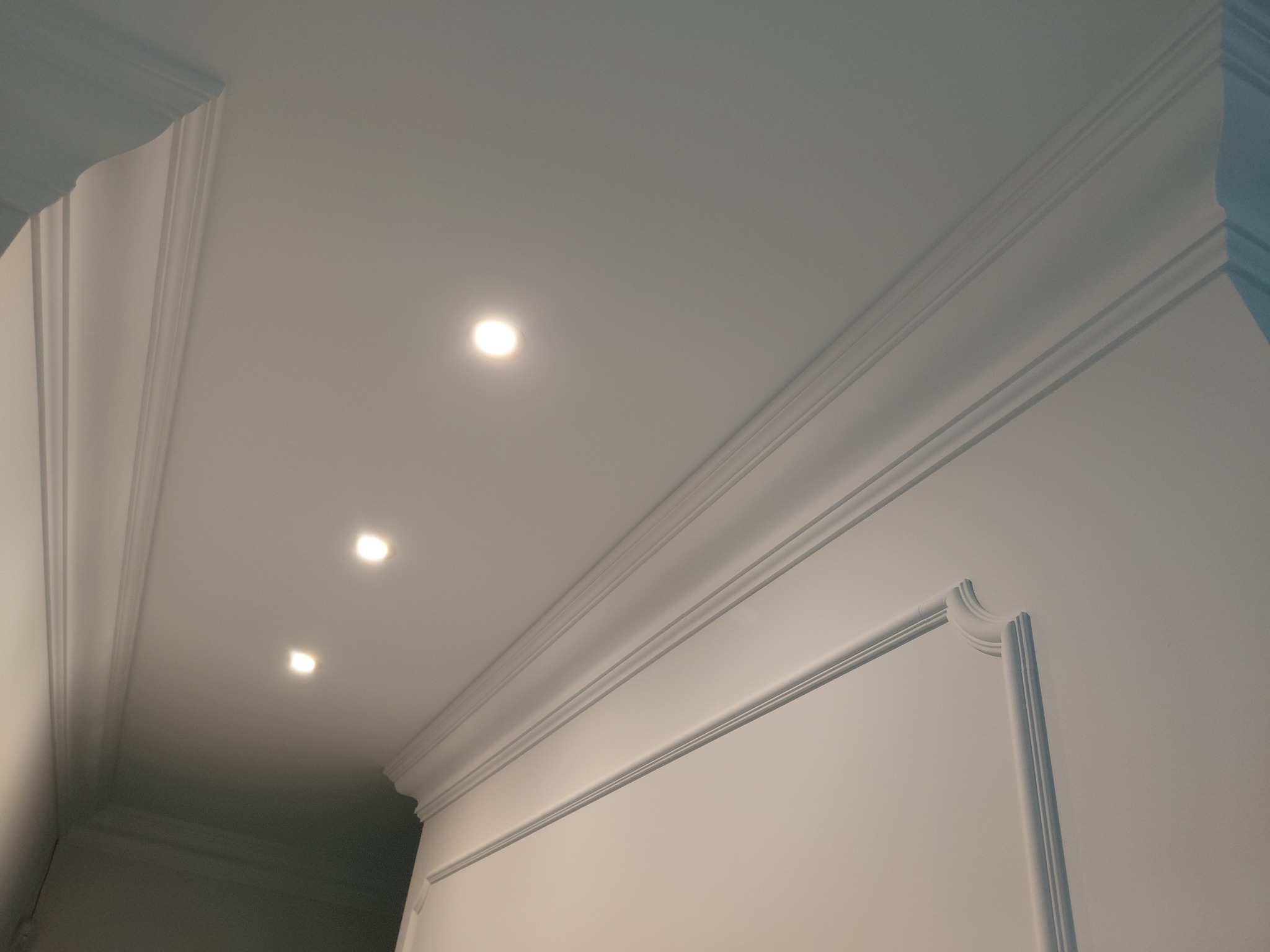 Białe, eleganckie wykończenie sufitu z listwami dekoracyjnymi i trzema punktowymi lampami LED, widok z dołu, podkreślający detale sztukaterii.