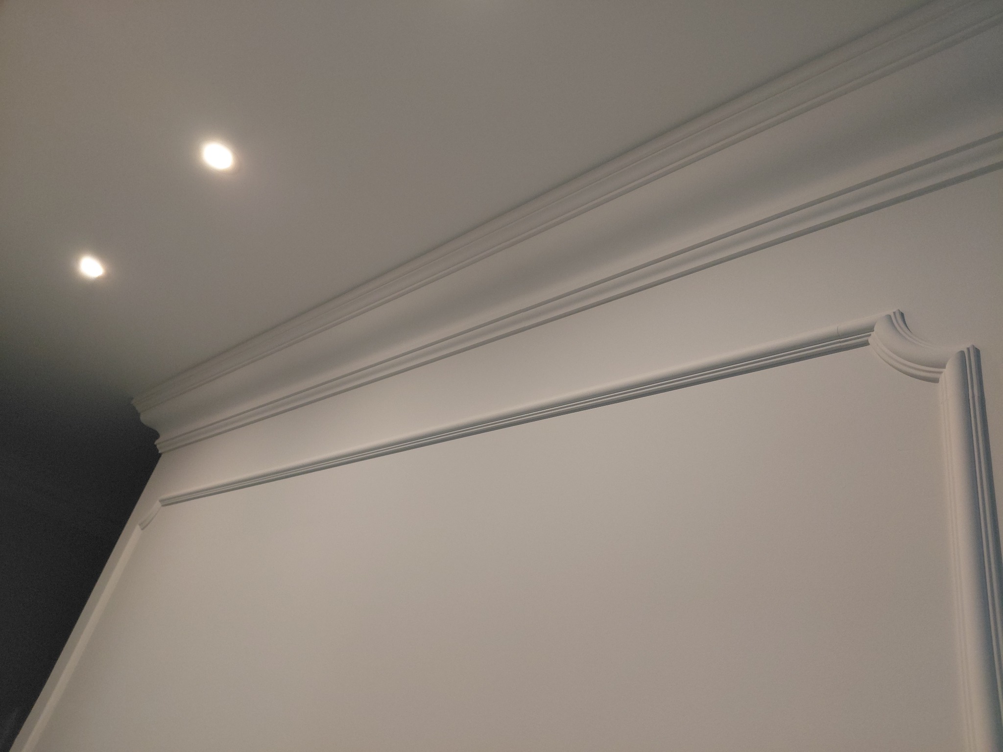 Biała ściana z dekoracyjnymi listwami przysufitowymi i ściennymi, oświetlona dwoma punktowymi lampami sufitowymi. Klasyczny detal wykończenia wnętrza.