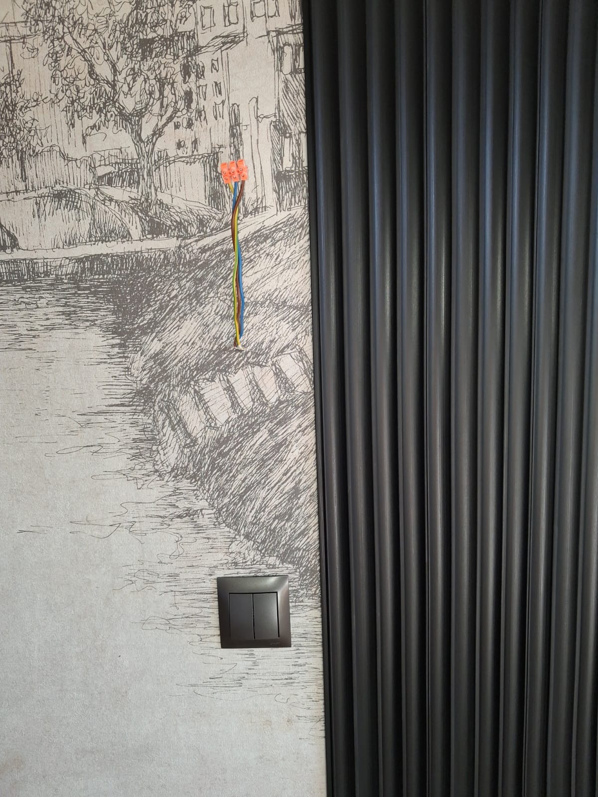 Szara ściana z dekoracyjną tapetą i panelem ściennym. Widoczne wystające przewody i włącznik światła. Etap prac wykończeniowych.