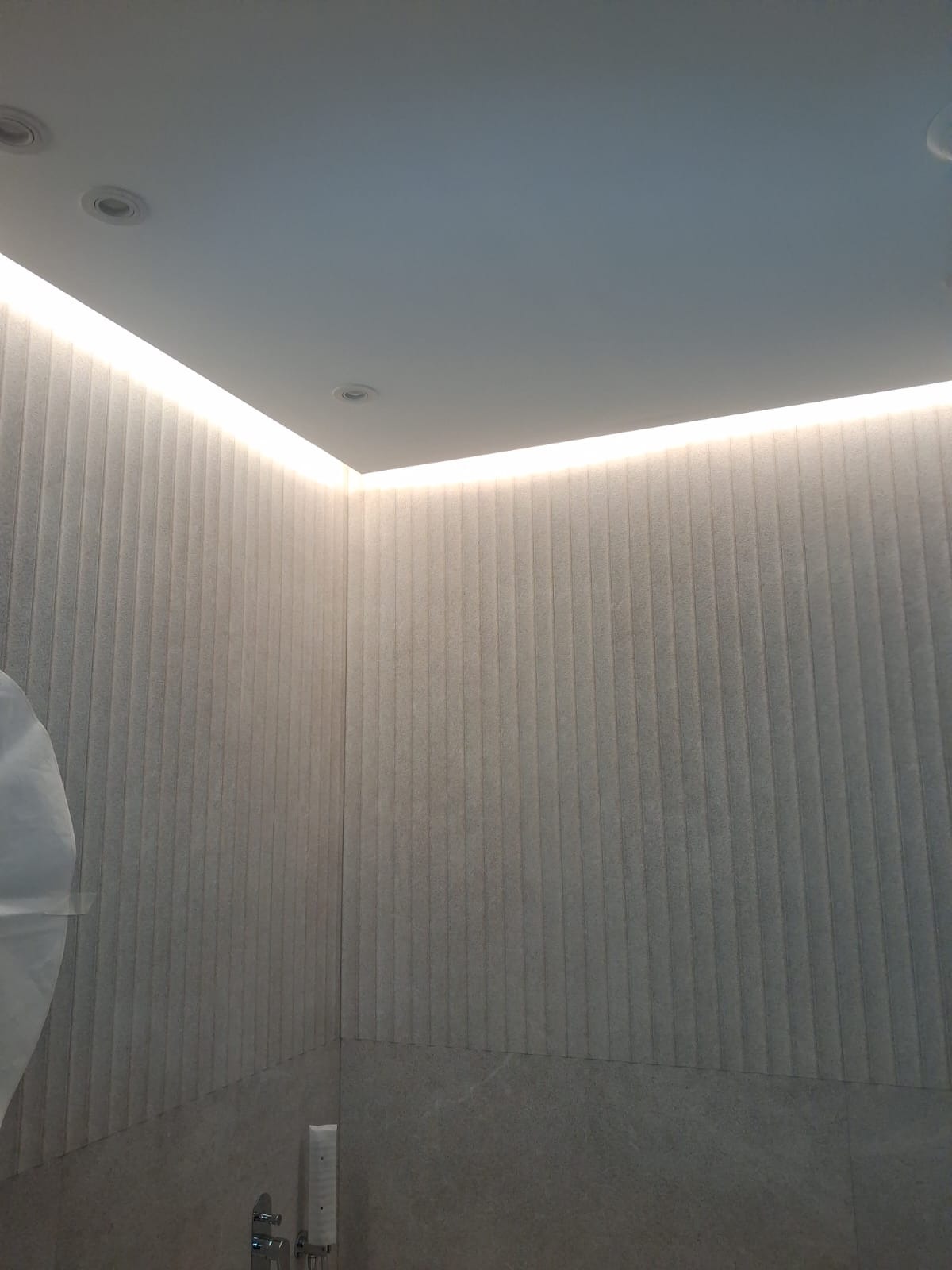 Nowoczesna łazienka z podświetlanym sufitem LED i panelami ściennymi w odcieniach szarości. Minimalistyczny design, widoczny fragment armatury.