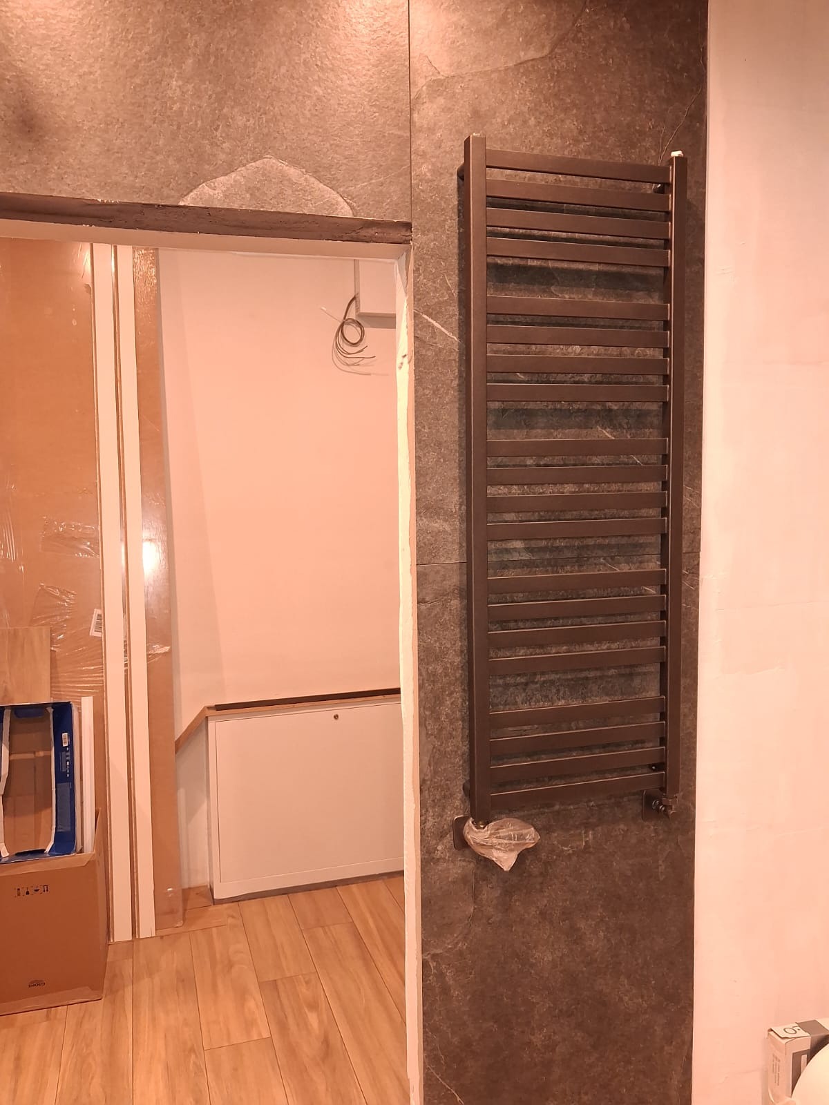 Zamontowany, brązowy grzejnik drabinkowy na szarej, kamiennej ścianie w łazience w trakcie remontu. Widoczne niedokończone wykończenia i materiały budowlane.