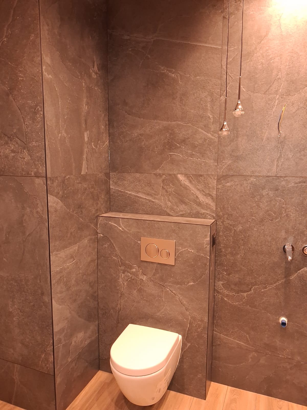Nowoczesna łazienka z brązowymi płytkami imitującymi kamień, podwieszana toaleta i minimalistyczne oświetlenie. Widoczne detale wykończenia i armatura.