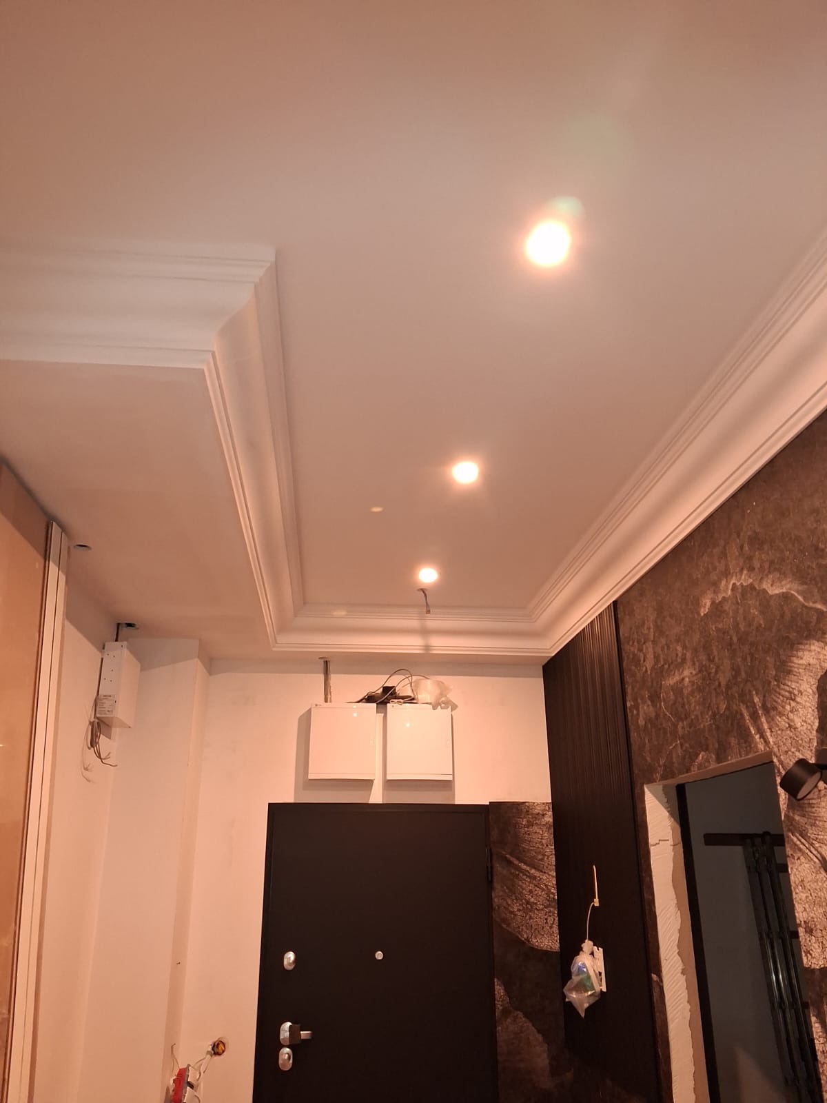 Wykończony sufit z listwami dekoracyjnymi i oświetleniem punktowym we Wrocławiu. Widoczna czarna ściana z wzorem i drzwi wejściowe. Detale wykończeniowe.