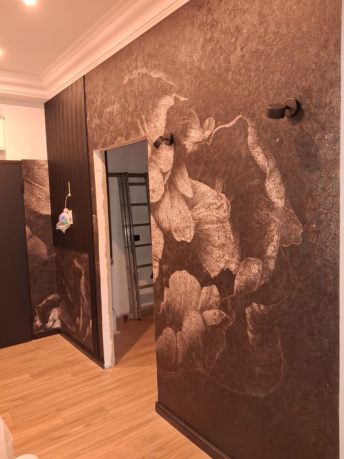 Wykończone wnętrze: brązowa ściana z motywem kwiatowym, drewniana podłoga i czarne, nowoczesne oświetlenie. Widoczny fragment drzwi i drabina w tle.