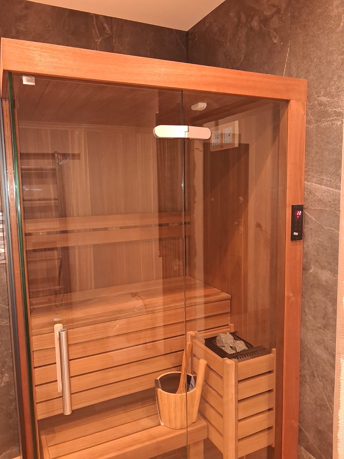 Nowoczesna sauna z drewnianym wykończeniem i szklanymi drzwiami, widoczny piec z kamieniami i drewniane akcesoria, całość wkomponowana w łazienkę z kamienną okładziną.