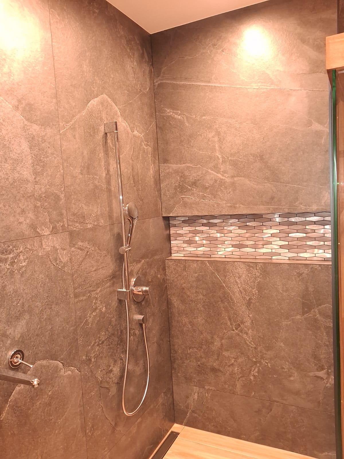 Wykończona kabina prysznicowa z szarymi płytkami imitującymi kamień i dekoracyjną wnęką z mozaiką. Chromowana armatura i odpływ liniowy.