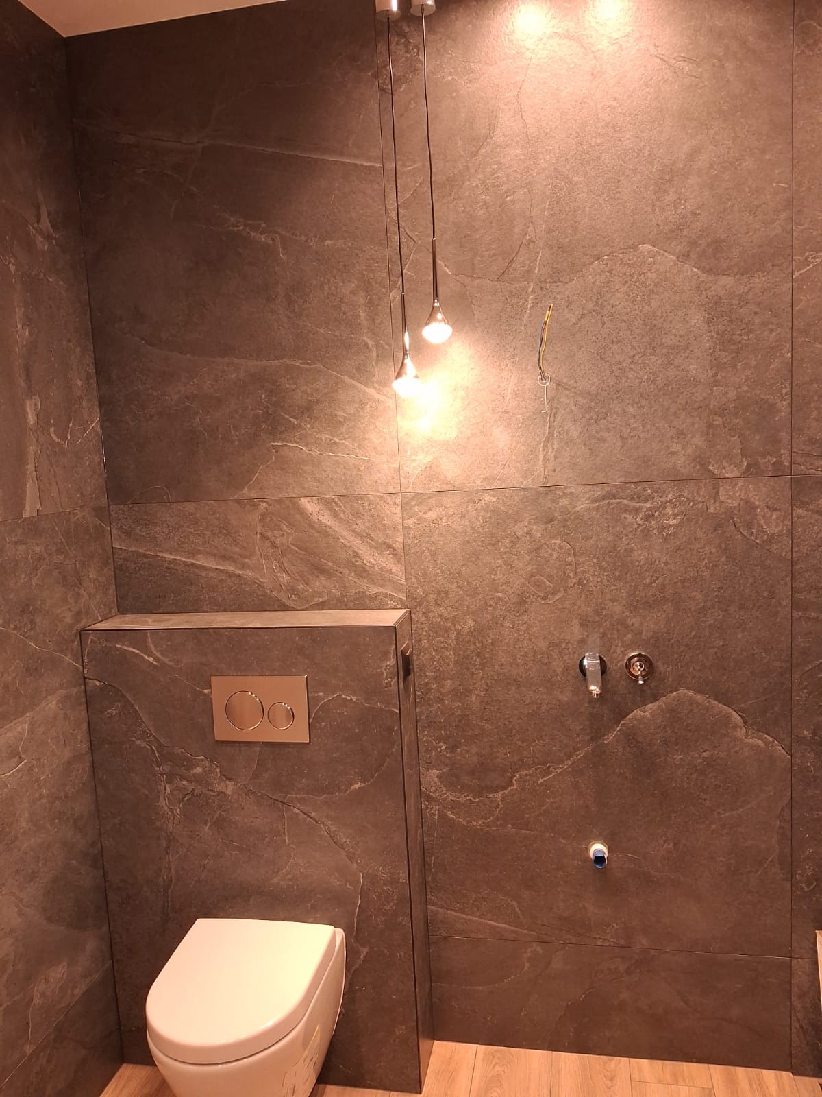 Wykończona łazienka z szarymi płytkami imitującymi kamień, podwieszana toaleta i minimalistyczne oświetlenie. Widoczne przyłącza wodne.